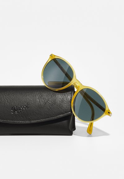 Lunettes de soleil jaunes en forme de œil de chat avec des verres verts et des branches fines, partiellement posées sur un étui en simili cuir noir avec un logo en relief.