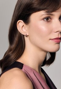 Srebrna kolczyk w kształcie obręczy noszony w lewym uchu, charakteryzujący się eleganckim designem. Osoba ma proste brązowe włosy i nosi bezrękawnik w kolorze mauve.