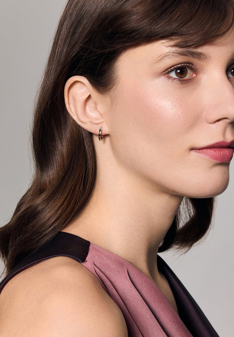 Srebrna kolczyk w kształcie obręczy noszony w lewym uchu, charakteryzujący się eleganckim designem. Osoba ma proste brązowe włosy i nosi bezrękawnik w kolorze mauve.