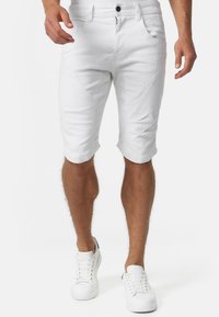 Short en jean blanc à coupe slim, avec cinq poches, une fermeture à bouton et des accents de couture discrets. Associé à des baskets blanches.