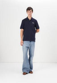 Marineblå polo med korte ermer, brodert logo; kombinert med lyseblå flared jeans og brune sko, stående mot en ensfarget bakgrunn.