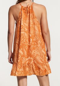 Woman wearing sleeveless orange dress with large floral print, halter neck tie, and ruffled hem, shown from the back.