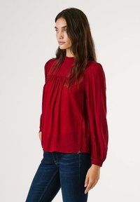 Blusa rossa, strutturata, a maniche lunghe con collo alto e dettagli arricciati. Abbinata a jeans scuri, presenta maniche elasticizzate e accenti sottili.