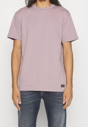 Osoba ubrana w prostą, jasnofioletową koszulkę z krótkim rękawem i niebieskie dżinsy stojąca na białym tle.