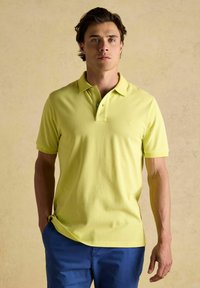 Limetkovozelená polo tričko z jemného materiálu s krátkymi rukávmi, trojbuttonovým uzáverom a malým vyšitým logom na hrudi. V kombinácii s modrými nohavicami.