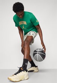 Camiseta de algodón verde con gráfico de "BOSTON", pantalones cortos estampados, calcetines negros de Jordan, zapatillas beige y un balón de baloncesto sobre un fondo gris.