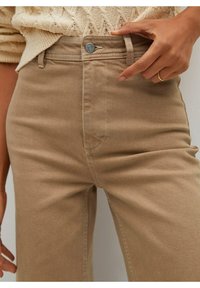 Bouton de fermeture des pantalons beiges taille haute, portés avec un pull en tricot crème, montrant une bague en or au doigt.