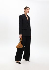 Čierny oblek na mieru s hlbokým výstrihom do V a zdobenými detailmi, skombinovaný s textúrovanou zlatou kabelkou a špicatými čiernymi lodičkami.