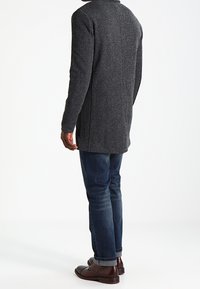 Mørkegrå lang frakke lavet af tekstureret stof, med høj krave, lige snit og sidesøm. Kombineret med blå jeans og brune sko.