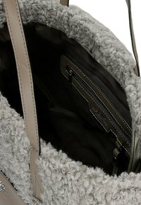Zbliżenie beżowej torby na ramię z zewnętrzną powłoką z kręconego futra, skórzanymi uchwytami i ciemnym wnętrzem z tkaniny, zawierającym kieszeń na zamek.