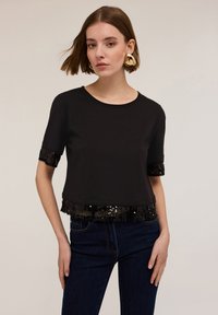 Camiseta negra recortada de manga corta, con un dobladillo y acentos de manga con lentejuelas, hecha de una tela suave. Combinada con jeans oscuros.