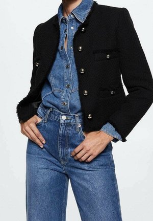 Femme portant une veste courte noire texturée avec des boutons métalliques par-dessus une chemise en denim bleu rentrée dans un jean taille haute bleu.