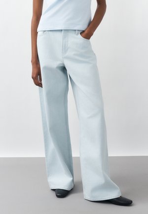 Osoba v svetlomodrých džínsach s širokými nohavicami, s jednou rukou v vrecku a čiernymi topánkami so štvorcovou špičkou na jednoliatom pozadí.