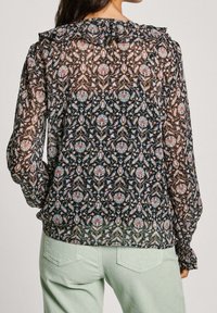 Doorzichtige zwarte blouse met bloemenpatroon in tinten roze en blauw, met een gerimpelde halslijn en geplooide mouwen, gecombineerd met lichtgroene broeken.