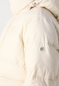 Puffet cremede jakke med quiltet design, der har en logo-knap og en tæt hætte for ekstra varme. Letvægts stofstruktur.