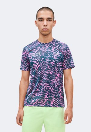 Νέος άνδρας με κοντά μαλλιά που φορά ροζ και σκούρο μπλε πουκάμισο με κοντά μανίκια και σορτς ανοιχτού πράσινου χρώματος, κοιτάζει μπροστά.