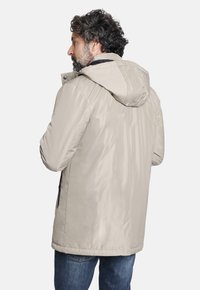 Chaqueta beige con capucha y un acabado suave y resistente al agua. Presenta bolsillos laterales con cremallera, puños elásticos y una silueta ligeramente ajustada.