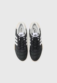 Black athletic shoes with a quilted design, three white stripes, gum sole, and a suede accent at the heel. Black laces and inner lining.