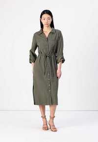 Vestido verde-oliva com botão, colarinho, mangas longas arregaçadas nos punhos, cinto de nó na cintura e detalhes de fendas laterais.