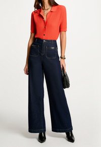 Czerwona koszula o fakturze, z krótkim rękawem i guzikami, zestawiona z szerokimi spodniami z ciemnego denimu, posiadającymi przednie kieszenie i widoczne przeszycia.