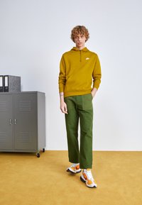 Sweatshirt mostarda da Nike com bolso canguru, calças largas verdes e sapatilhas multicoloridas. Armário cinza ao fundo. Chão amarelo.