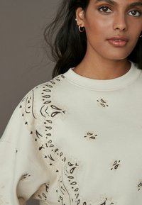 Light beige sweatshirt featuring intricate black and brown decorative patterns along the neckline and sleeves. Soft, smooth texture.