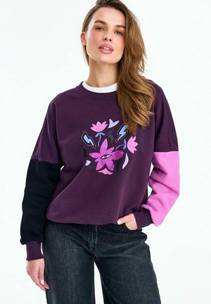 Kobieta w fioletowej bluzie z czarnymi i różowymi rękawami, z centralnym wzorem kwiatów, serc i błyskawic.