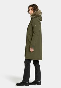 Olijfgroene parka met een met bont gevoerde capuchon, een rits aan de voorkant en zijzakken; gecombineerd met zwarte broek en schoenen. Zachte textuur, knielange ontwerp.