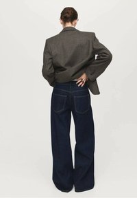 Blazer de lana gris sobre jeans de pierna ancha azul oscuro, con bolsillos traseros visibles y costuras amarillas contrastantes. Diseño minimalista y textura suave.