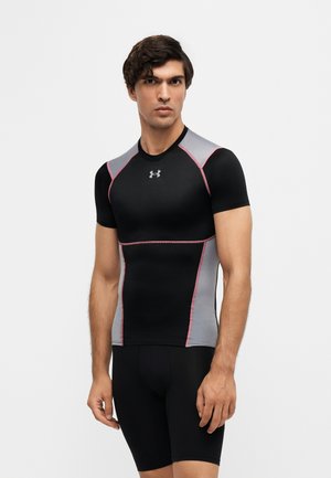 Homme portant un t-shirt de compression noir et gris à manches courtes et un short noir, debout devant un fond blanc uni.