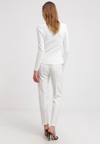 Vrouw in een wit, gestructureerd longsleeve shirt en witte op maat gemaakte broek, zonder schoenen op een witte achtergrond, met haar rug naar de camera gekeerd.