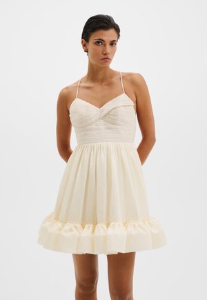 Femme aux cheveux courts et foncés portant une mini robe crème sans manches avec fines bretelles, un corsage plissé et un ourlet à volants.
