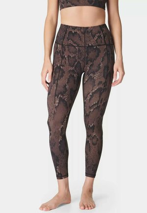 Vrouw draagt een high-waisted legging met bruin-zwart slangenprint en een bijpassend cropped topje, staat blootsvoets tegen een witte achtergrond.