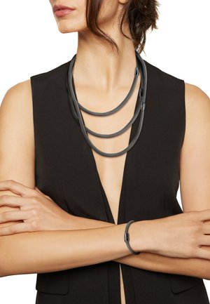 Grå fletning halskæde med tre strenge, der har slanke sorte hardware-detaljer. Matchende grå armbånd med en strømlinet lås.