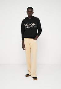 Μαύρη κουκούλα φούτερ με λευκό λογότυπο σε σενάριο, συνδυασμένη με ανοιχτό μπεζ παντελόνι σε φαρδιά γραμμή και μαύρα παπούτσια με λαστιχένια σόλα καφέ.