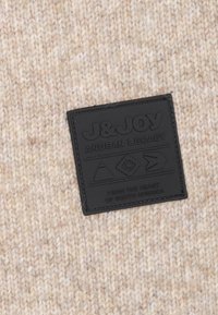 Gebreide stof in lichte beige tinten met een gestructureerd patroon, voorzien van een zwart rechthoekig leren label met de opdruk "J&JOY" en geometrische iconen.