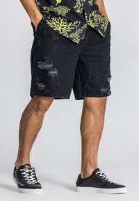 Shorts de mezclilla negra con detalles desgastados y bordes deshilachados, con un corte relajado. Combinados con zapatillas negras de suela blanca.