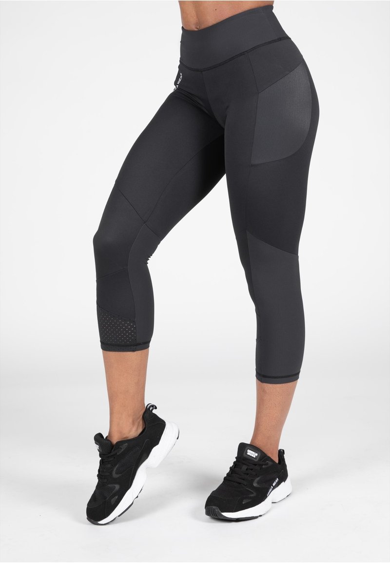 Zwarte capri-legging met een hoge tailleband, voorzien van textuurpanelen en geperforeerde accenten aan de onderkant van het been. Gecombineerd met zwarte sportschoenen.