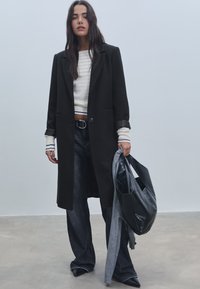 Musta villa takki, jossa on räätälöity muotoilu, harmaa teksturoitu neule, tummat farkut ja musta nahkareppu. Sisältää leveät hihat ja napitus.