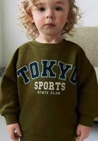 Olivově zelená mikina s velkým grafickým nápisem "TOKYO SPORTS" v námořnické modré a bílé barvě, s žebrovanými manžetami a kulatým výstřihem. Měkká textilie.