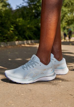 Persona de piel oscura que lleva zapatillas de correr gris claro con suelas blancas, de pie en un sendero al aire libre iluminado por el sol, con vegetación verde de fondo.