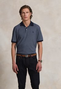 Camisa polo azul-marinho com um padrão, gola escura e mangas curtas, combinada com jeans escuros e um cinto castanho.