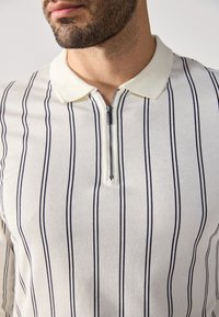 Polo shirt zippé blanc avec des rayures verticales bleu marine, un col doux et un détail à fermeture éclair argenté au niveau du cou. Tissu texturé.