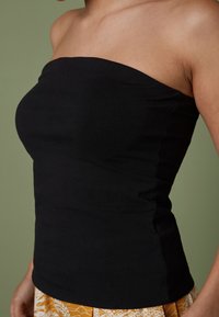 Haut sans bretelles noir en tissu extensible, avec un design épuré et une silhouette ajustée. L'arrière-plan est d'un vert doux.