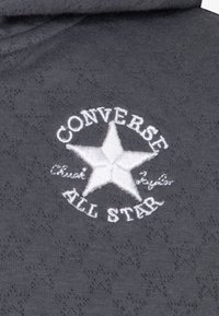 Sivi materijal s suptilnim uzorkom sadrži izvezenu bijelu zvjezdicu i tekst "CONVERSE ALL STAR" oko nje.