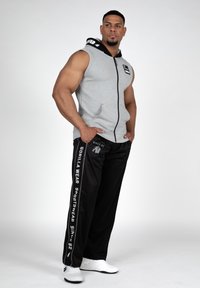 Grijze mouwloze hoodie met een zwarte capuchon en rits, gecombineerd met zwarte sweatpants met witte zijstrepen en een logo. Getextureerde stof.