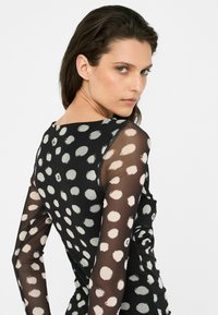Maglietta a maniche lunghe con design a pois bianchi e neri, caratterizzata da un tessuto trasparente sulle maniche e un corpo nero opaco. Texture liscia, forma aderente.