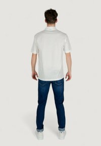 Camicia bianca a maniche corte con tessuto strutturato e colletto, abbinata a jeans in denim blu scuro e sneakers bianche. Vista posteriore.