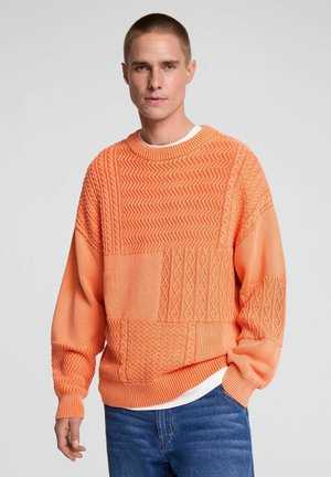 Uomo che indossa un maglione arancione strutturato sopra una camicia bianca e jeans blu, in piedi con una mano in tasca su uno sfondo semplice.