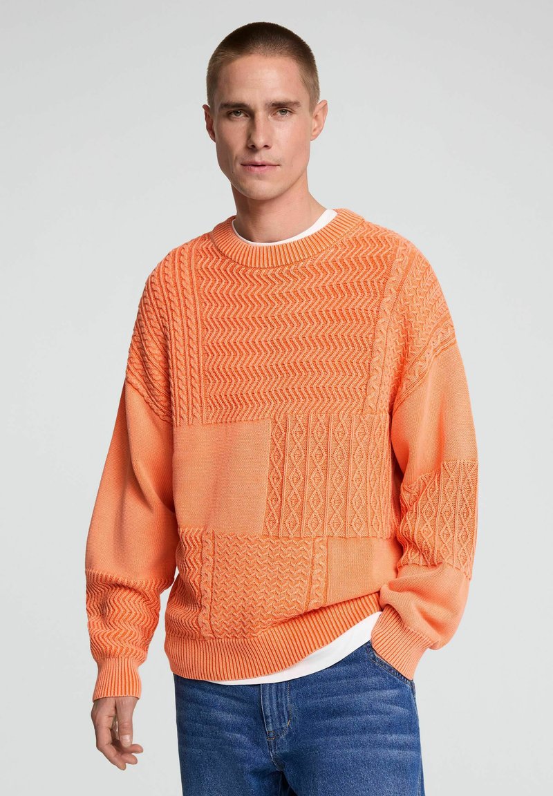 Homme portant un pull orange texturé sur une chemise blanche et un jean bleu, debout avec une main dans la poche devant un fond uni.